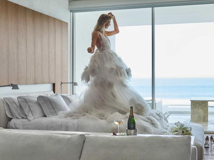 A bride poses in her wedding gown while standing on the bed of an oceanside hotel room