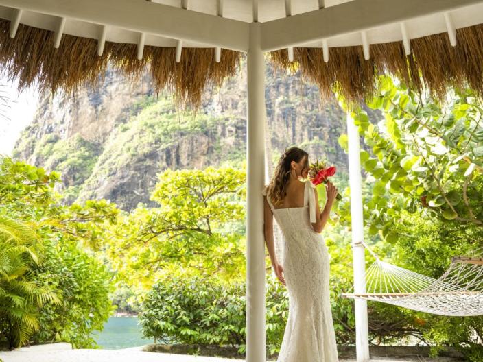 A bride smells her bouquet in a courtyard, next to a hammock looking out on a tropical landscape