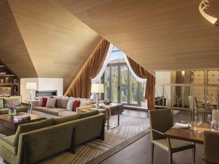 The Viceroy Kopaonik Serbia Penthouse Suite common area, with an L-shaped couch, large book case, vaulted ceilings, a writing desk and a window looking out on the ski slope in summer