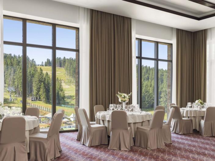 The Aero room at Viceroy Kopaonik Serbia, with padded chairs surrounding round banquet tables on a pink striped carpet, and large windows looking out on a ski slope in summer time