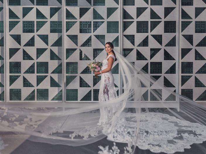 A bride poses with her bouquet next to a concrete mosaic wall