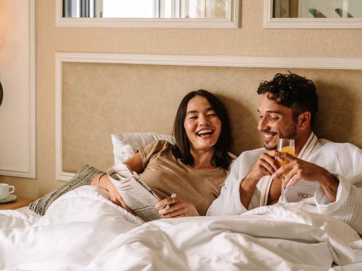 A couple laying in bed with the paper and a mimosa