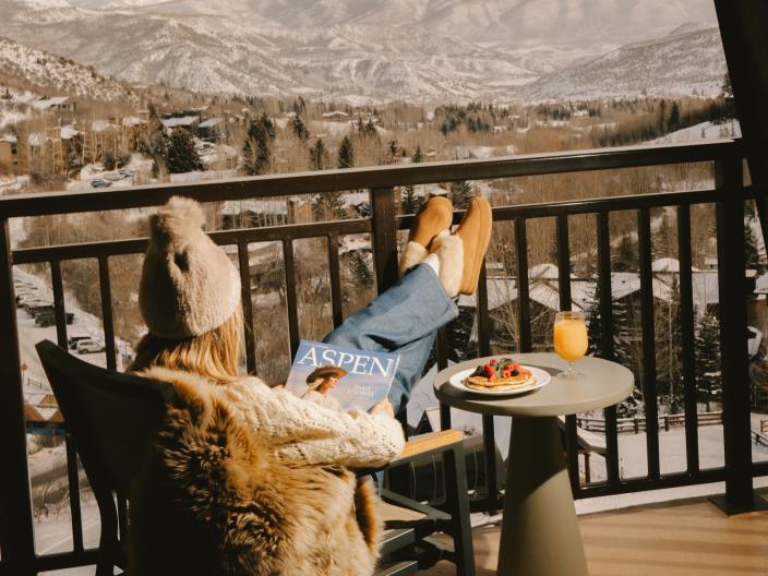 A woman reads a magazine with her feet up, seated in a chair on a balcony overlooking a snowy Colorado mountain range