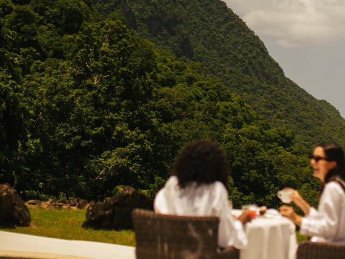 Two diners eat on a terrace at the base of a mountain