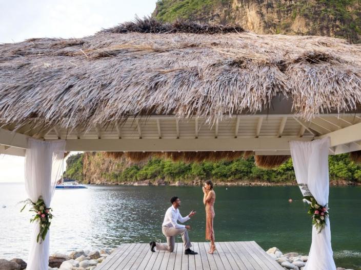 A man proposing on one knee to his fiancee, in an outdoor straw-roof gazebo at the base of the Pitons at Viceroy Sugar Beach