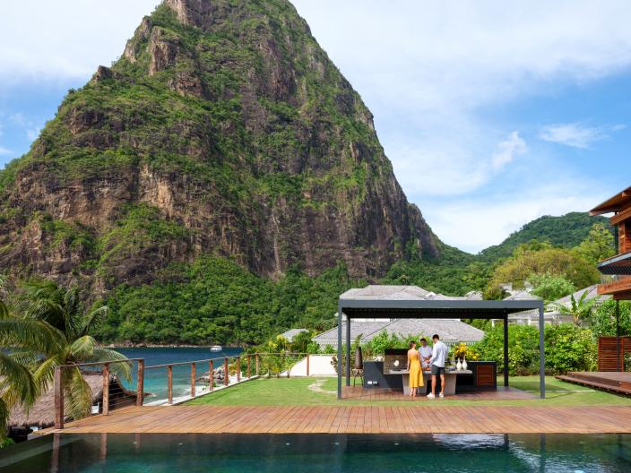 Two guests get served a meal from an outdoor hut, with a cove at the foot of a large tree-lined mountain in the background
