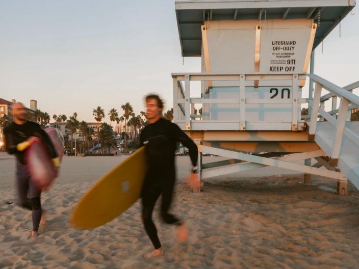 two people in wetsuits carrying longboards run in front of lifeguard station at beach