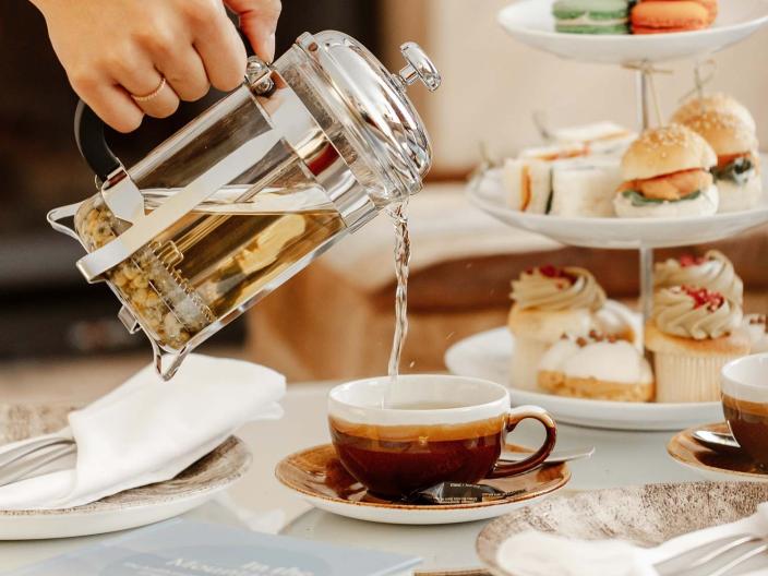 A person pours tea next to a tea tower with sandwiches and macarons