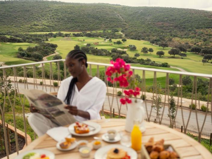 A woman reads a newspaper on a Viceroy Ombria Algarve patio, with in-room breakfast in front of her and the rolling green Algarve hills behind her.