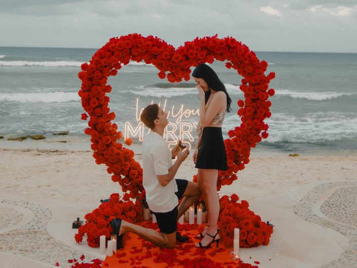 person on their knee with another person standing on beach with red flowers in heart shape