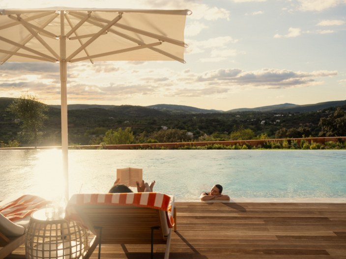 A person sits in a deck chair reading, while their partner lounges in the pool.