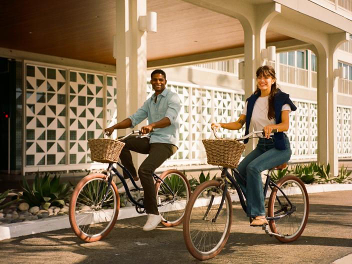 Um casal sorridente passa de bicicleta cruiser a condizer, com cestos de vime, em frente à fachada de estilo moderno de meados do século de um hotel chamado «VICEROY».