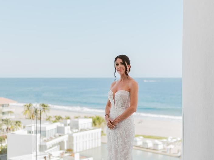 Bride in a white strapless lace wedding gown stands on a balcony overlooking a bright blue ocean, white resort buildings, and a beach below.