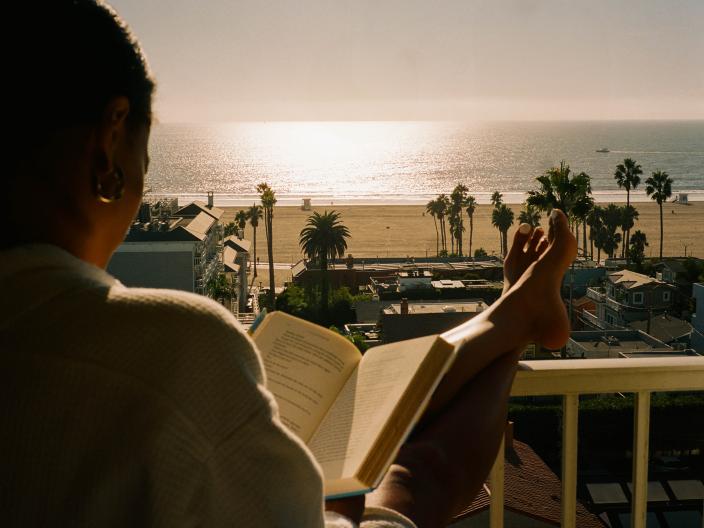 Una mujer vestida con una túnica blanca lee un libro abierto mientras descansa en un balcón con vistas a una playa bañada por el sol y rodeada de palmeras.
