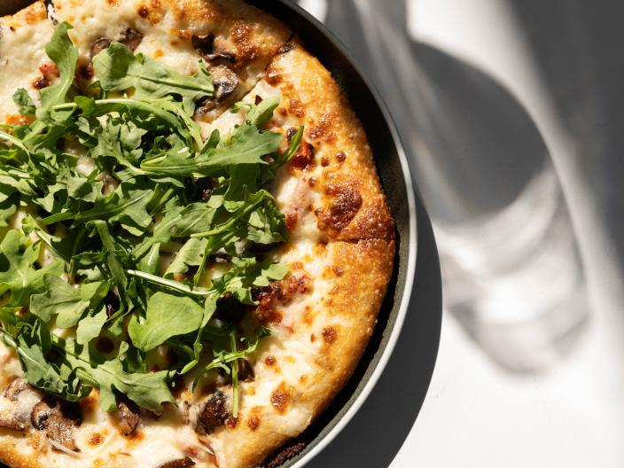 Primer plano desde arriba de una pizza redonda con salsa blanca, champiñones y rúcula fresca, servida en un plato oscuro sobre una superficie blanca brillante, con espectaculares sombras creadas por la luz del sol.