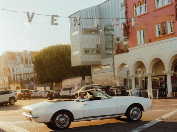 Un descapotable vintage blanco recorre una calle bajo el letrero de «VENICE», con un edificio rosa al fondo, durante la hora dorada.