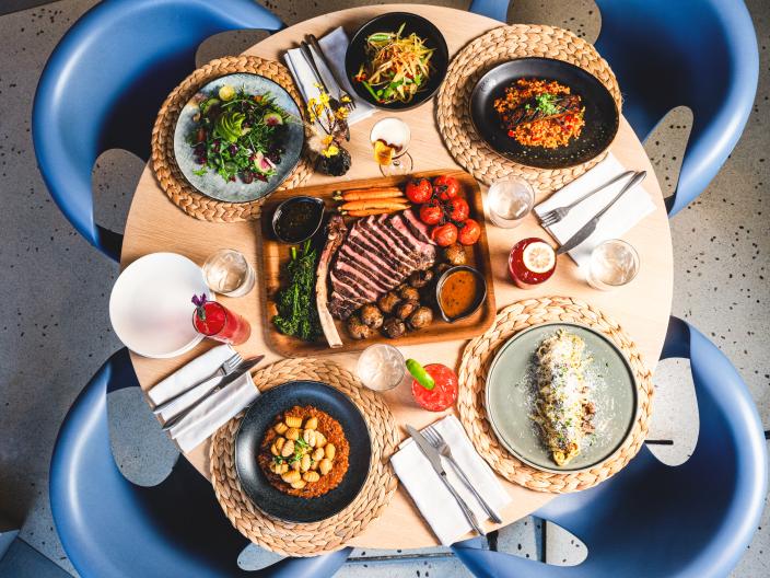 Vista aérea de uma mesa de jantar de madeira com um grande prato de bife fatiado, vários acompanhamentos, cocktails e cadeiras azuis modernas.