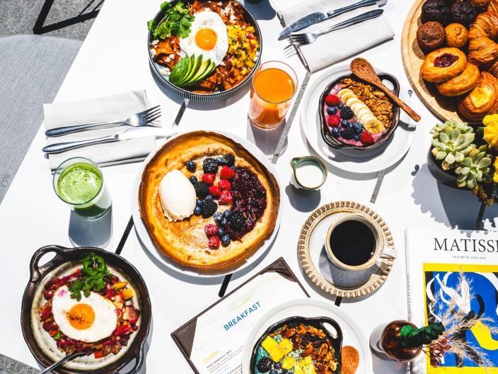Vista aérea de uma mesa ao ar livre bem iluminada, repleta de diversos pratos de brunch, incluindo ovos, panquecas, taças de granola, pastéis, café e sumos.