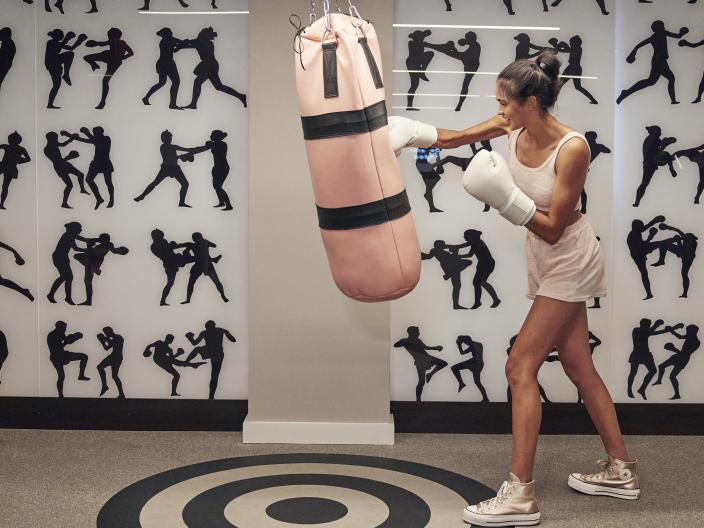 Une femme portant des gants de boxe frappe un sac de frappe rose devant un mur orné de silhouettes représentant des arts martiaux.