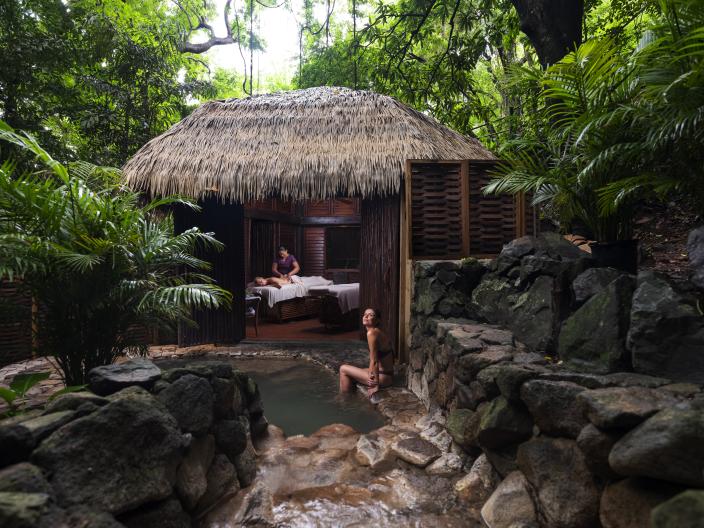A woman sits at the edge of a natural hot spring next to a thatched-roof spa where a massage is being given.