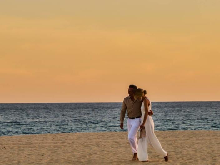 Un couple se promène bras dessus bras dessous sur une plage de sable, sous un coucher de soleil chaleureux.