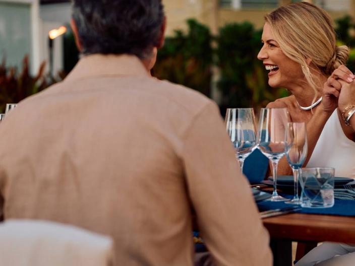 Une femme rit, assise à une table avec un homme ; sur la table se trouvent des verres à vin et des couverts.