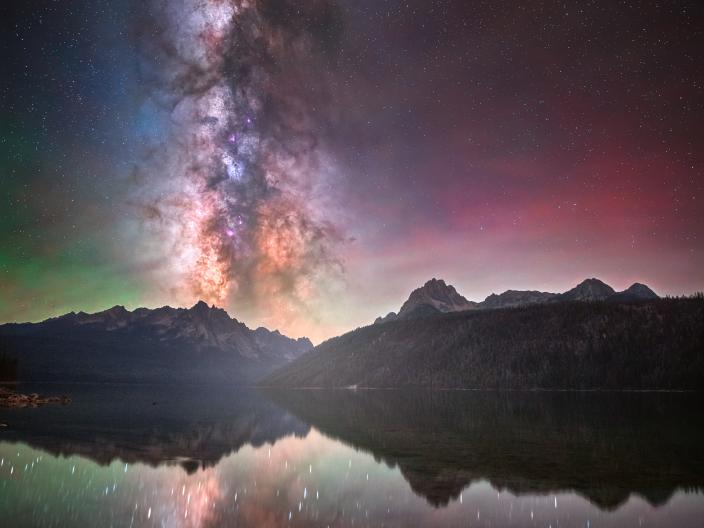 A silhueta de montanhas escarpadas e de um pinhal refletida num lago tranquilo, sob uma Via Láctea cintilante e uma aurora ténue.