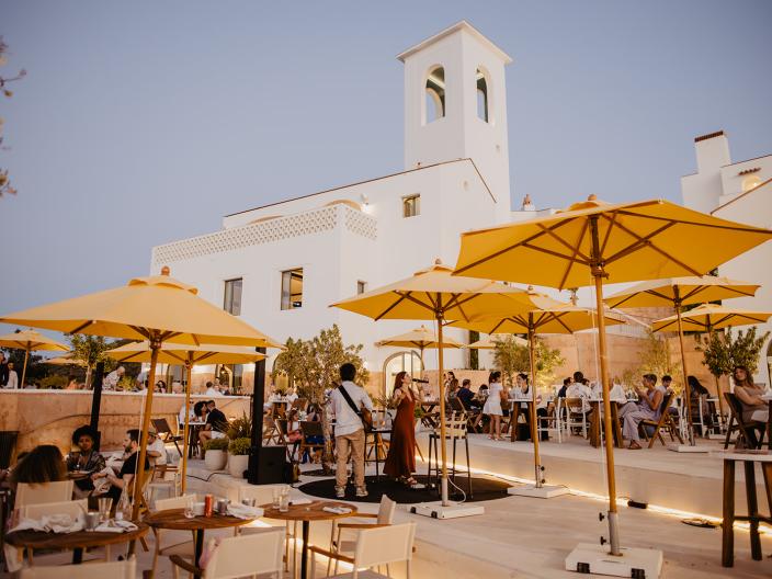 Outdoor café terrace filled with diners beneath yellow umbrellas, featuring a white Mediterranean-style building and bell tower at dusk.
