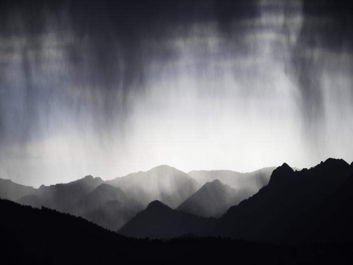 Listras dramáticas de chuva a cair sobre cumes montanhosos em camadas, recortados contra o céu, sob uma luz evocativa.