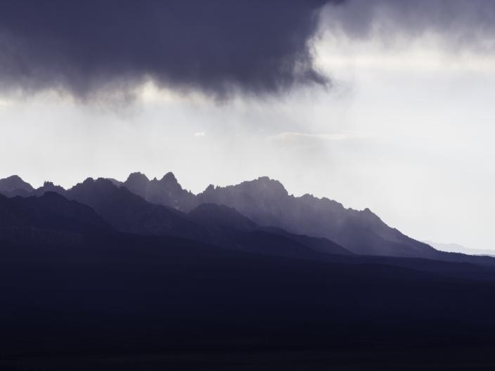 Uma nuvem negra de tempestade paira sobre os picos montanhosos irregulares e distantes, com sutis feixes de luz.