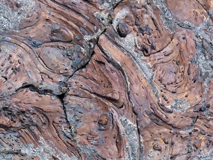 Imagem em grande plano da textura de uma rocha de lava pahoehoe castanho-avermelhada, desgastada pelo tempo e com um padrão espiralado, apresentando depósitos minerais arenosos.