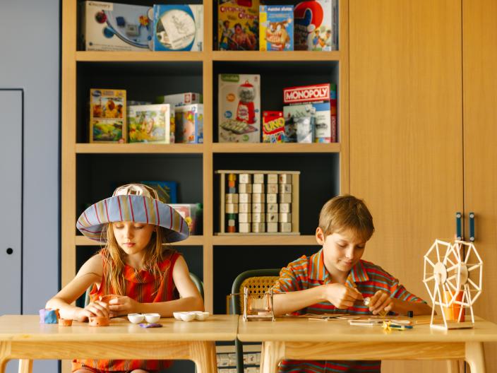 Two children with crafting supplies at a wooden table in front of a bookshelf filled with games and toys.
