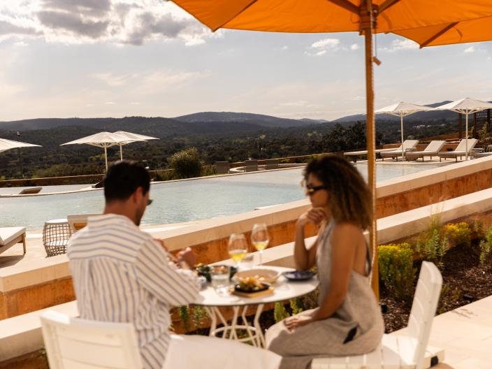 A couple dines outdoors by an infinity pool with a scenic mountainous landscape.