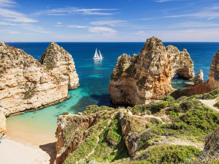 A sailboat navigates turquoise waters between dramatic sea cliffs and a small sandy beach.
