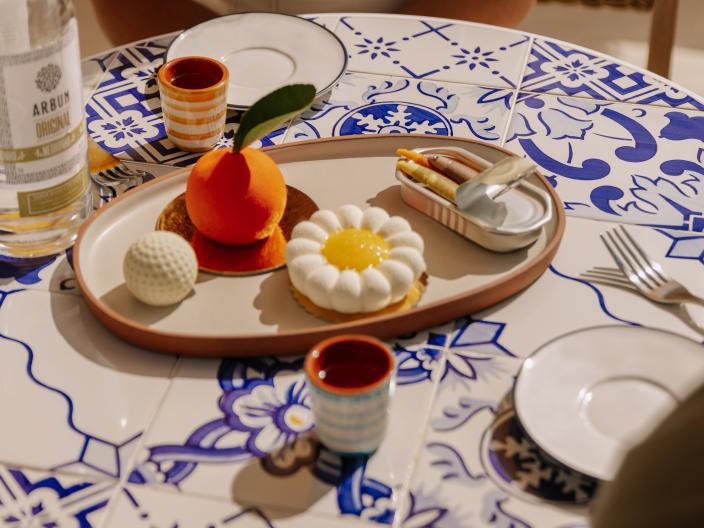 Dessert platter with orange-shaped mousse and floral cake served on a tiled table with drinks.