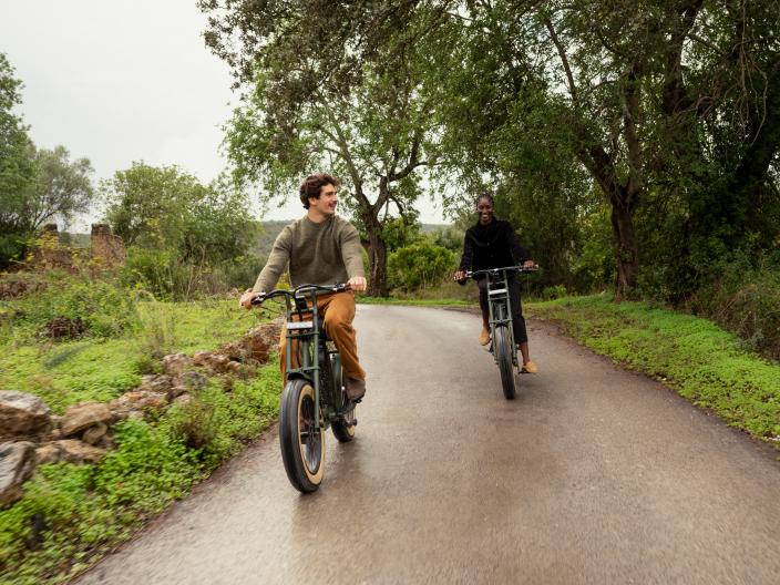Two people ride electric bikes on a wet, paved road lined with greenery and trees.