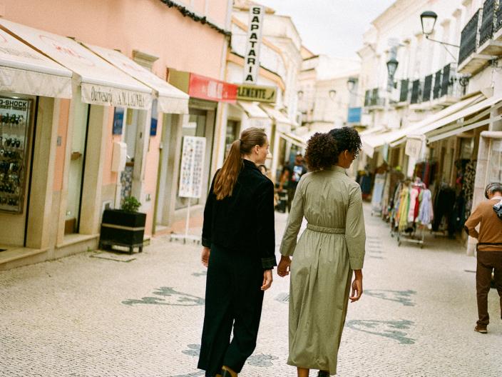 Two women walking away from the camera on a cobblestone street lined with pastel-colored buildings and shops in a European town.