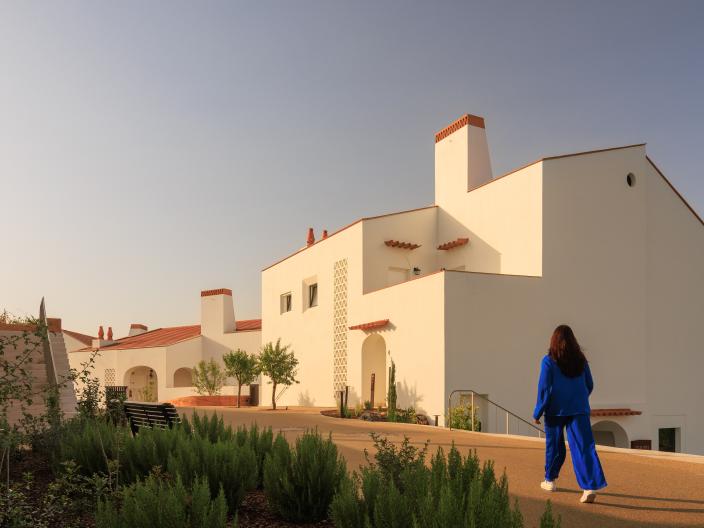 A woman in bright blue clothing walks along a path toward white, modern buildings with red-tiled roofs under a clear sky.