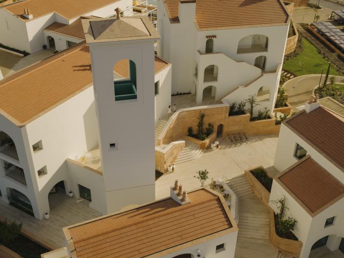 Aerial view of a modern resort with white stucco buildings and terracotta roofs, featuring an ornate tower and tiered courtyards.