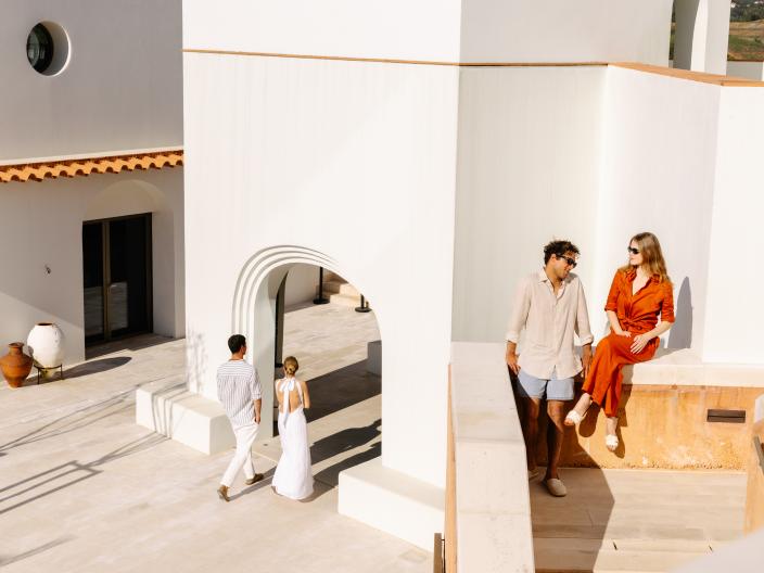 Couple in orange and beige outfits sit on a sunlit white terrace while two people walk beneath an arched courtyard entrance.