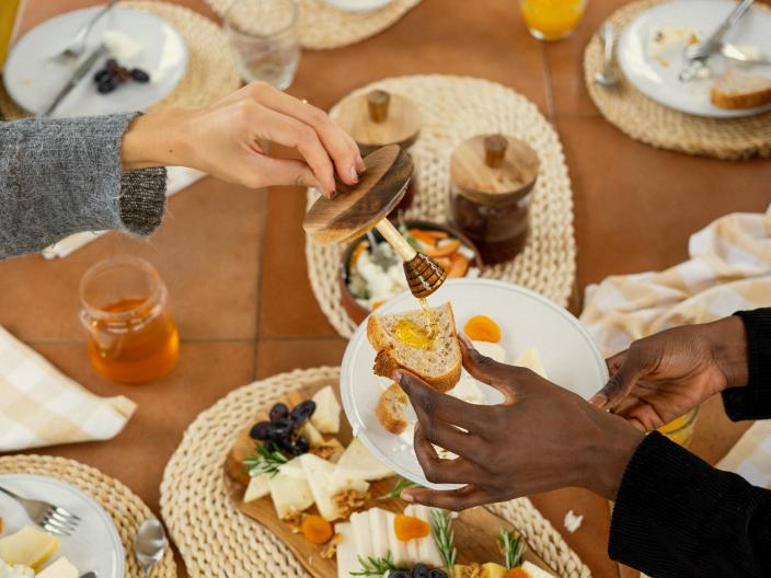 People share a brunch table with toast, honey, orange juice, yogurt, fruit, cheese, and charcuterie served on woven placemats.