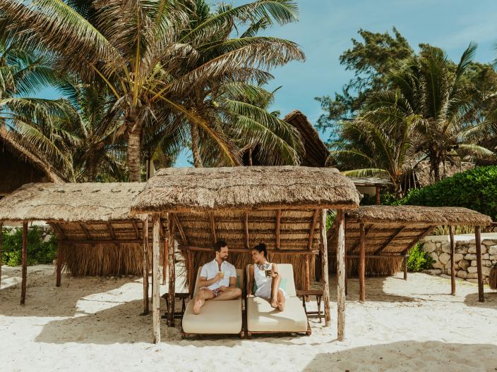 A couple relaxes under a thatched cabana on a white-sand tropical beach surrounded by palm trees.