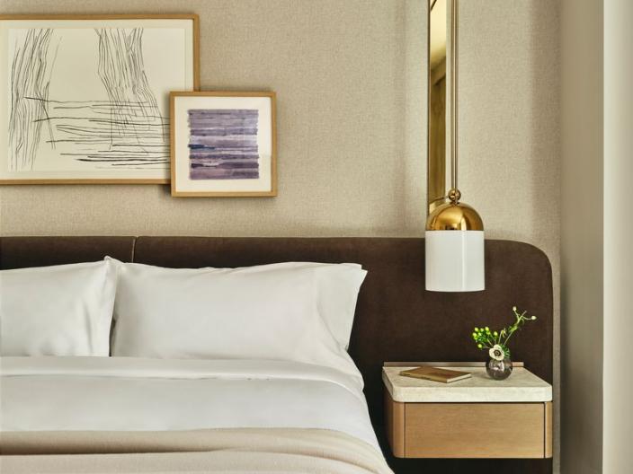 A neatly made bed with white pillows and beige bedding beside a wooden nightstand, wall lamp, flowers, and framed artwork.