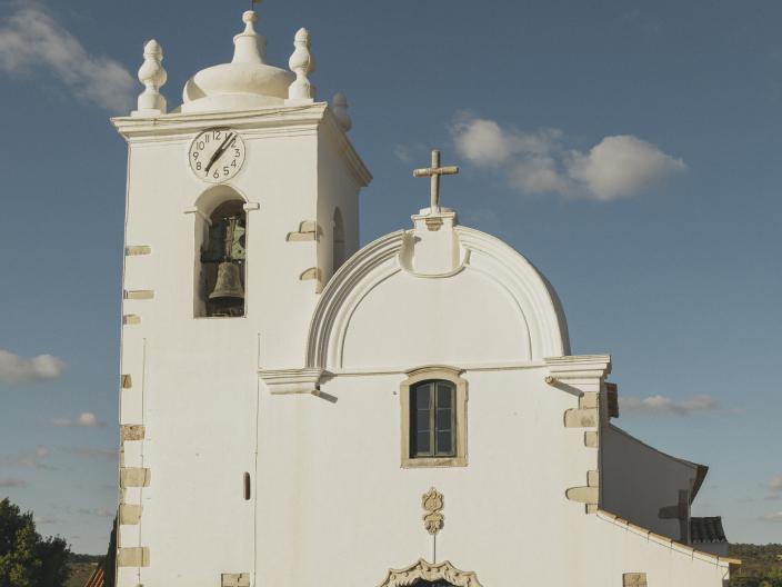 White historic church with a clock tower, domed pinnacles, ornate arched entrance, and cross-topped façade under a blue sky.