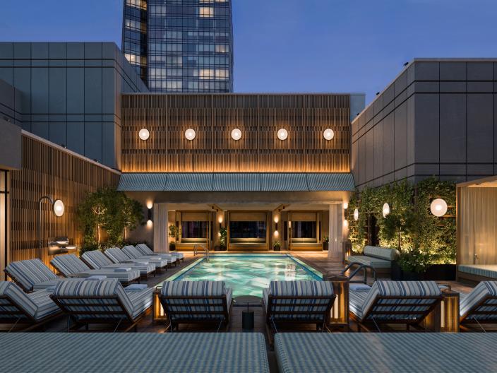 A rooftop swimming pool at dusk is lined with striped lounge chairs, with modern buildings, warm lights, and a glass tower behind it.
