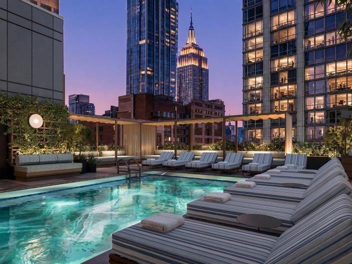 Une piscine sur le toit, bordée de transats rayés, donne sur les gratte-ciel illuminés de New York au crépuscule.