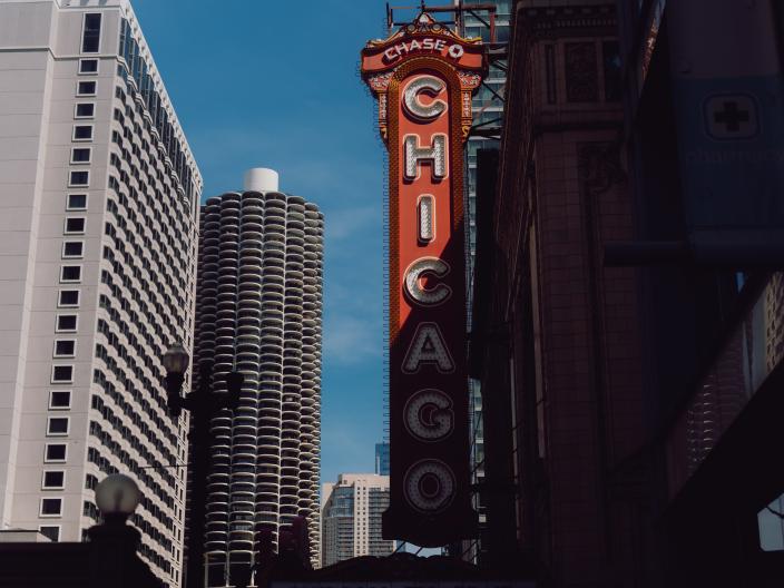 Um letreiro vertical vermelho de teatro com as inscrições «CHICAGO» e «CHASE» ergue-se entre os edifícios do centro da cidade, incluindo uma torre redonda composta por cilindros empilhados.