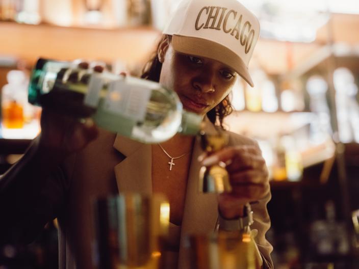 Uma mulher atrás do balcão do bar usa um boné com a inscrição «CHICAGO» e um colar com uma cruz, enquanto segura copos perto da câmara.