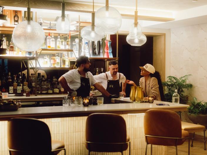 Três pessoas conversam no balcão de um bar, repleto de garrafas, copos de latão e bancos, sob candeeiros de vidro em forma de globo suspensos no teto.