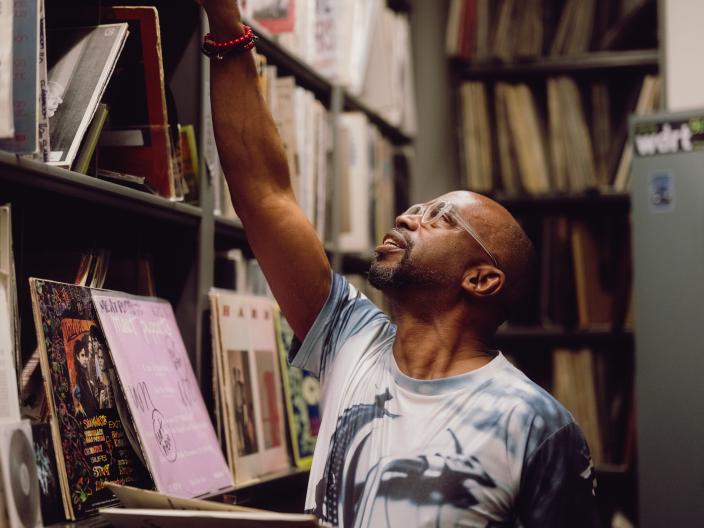 Um homem de óculos estica o braço para pegar num disco numa prateleira alta numa loja de música, enquanto segura uma capa de disco aberta.